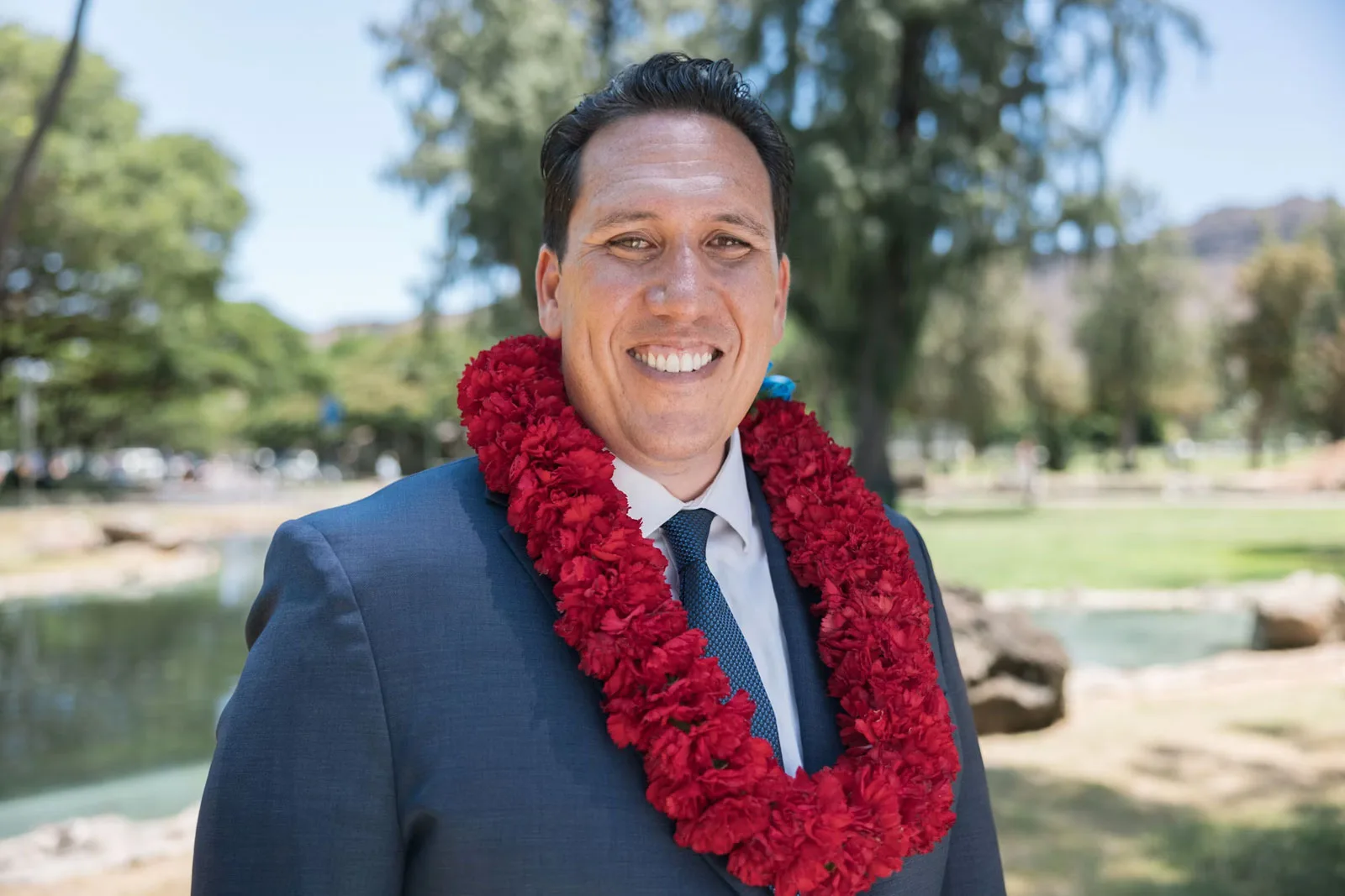 Jarrett Keohokalole smiling outdoors in a suit, wearing a red carnation lei, with a park pond and trees in the background