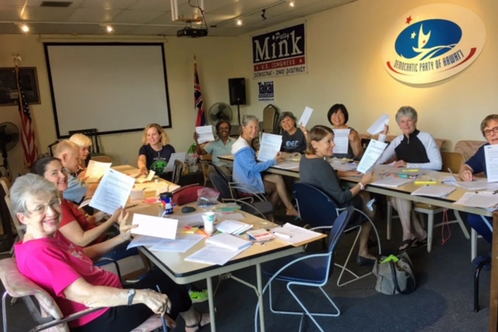 3BEE031B-2762-4B6F-AF4F-EBE77CB6DA75 Group of people posing with their postcards at the weekly postcarding party in Kailua, hosted by the Blue Wave and Indivisible Windward Oahu.