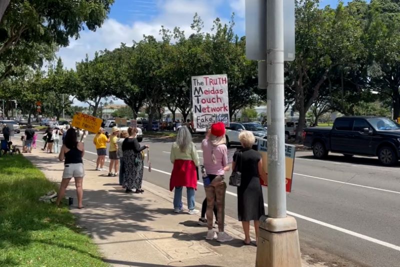 People gathered along a busy street in West Oahu as cars drove by during the No Dictators Protest in October 2026, organized by Indivisible to advocate for democracy.