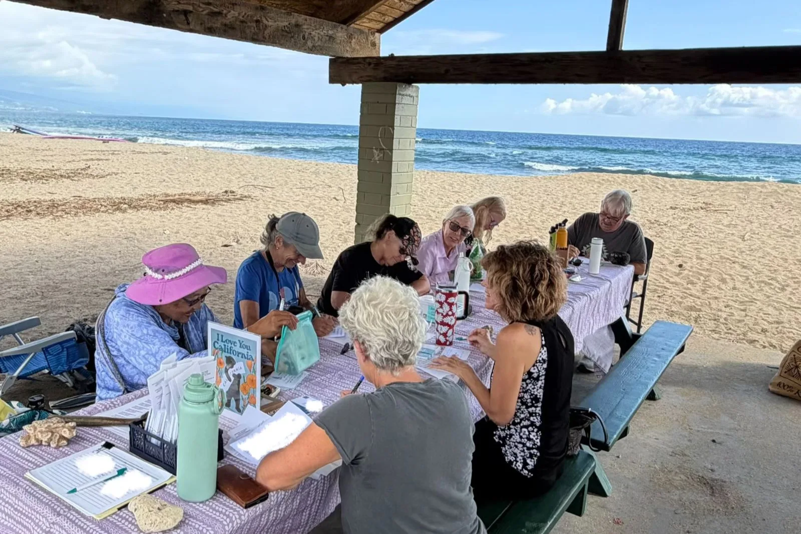 kona chapter leadership-1600x1067 People laughing while at a barbecue table writing postcards with the sand and the Pacific Ocean in the background.